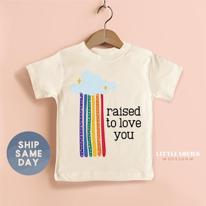 May include: Cream-colored toddler t-shirt with a rainbow design and the phrase "raised to love you" in black. The rainbow features a blue cloud, yellow stars, and colorful stripes. The shirt is hanging on a wooden hanger.