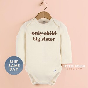 A white onesie with the text "only child big sister" printed on it, hanging on a wooden hanger against a peach background.