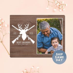 May include: A wooden photo frame with a picture of a man and a baby. The left side features a white graphic of a deer head, crossed rifles, and the text "BEST GRANDPA - BY A LONG SHOT". The frame is brown.