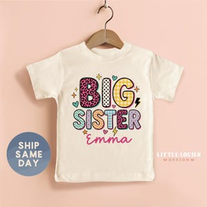 May include: A cream-colored short-sleeve t-shirt featuring the text BIG SISTER in colorful, patterned block letters including leopard print, gingham, and chevron designs. The name Emma is printed below in pink script, accented by small stars, hearts, and lightning bolts.