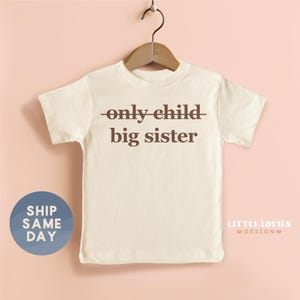 A white t-shirt with the text "only child big sister" printed on it, hanging on a wooden hanger against a pink background.