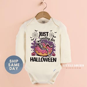 May include: Cream-colored baby bodysuit with long sleeves. The front features a cartoon skeleton relaxing on a pink donut, with the text "JUST waiting for HALLOWEEN". Includes spiderwebs, bats, and pumpkins.