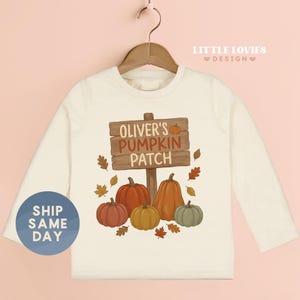 May include: A cream-colored long-sleeve shirt featuring a graphic of a wooden sign that reads "OLIVER'S PUMPKIN PATCH" with a small orange pumpkin icon. Below the sign are four pumpkins in orange, yellow, and green, surrounded by autumn leaves.