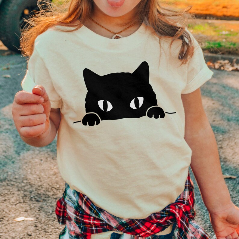 A person wearing a white t-shirt with a black cat graphic and pointing at the camera.