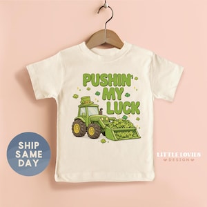 May include: A white t-shirt featuring a green tractor with a bucket full of shamrocks. The text "PUSHIN' MY LUCK" is printed above the tractor in green. The shirt is hanging on a wooden hanger.