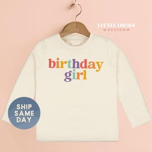 A white long-sleeved t-shirt with the text "Birthday girl" printed on it, hanging on a wooden hanger.