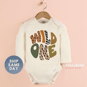 A white onesie with the text "WILD ONE" printed on it, hanging on a wooden hanger against a pink background.
