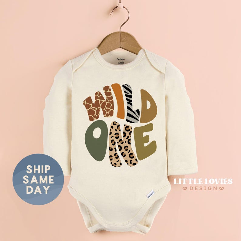 A white onesie with the text "WILD ONE" printed on it, hanging on a wooden hanger against a pink background.