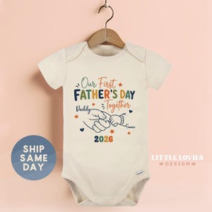 May include: Cream-colored baby onesie with the text "Our First Father's Day Together" in navy, green, and orange. The design includes a graphic of a parent's hand holding a baby's hand, with the name "Emma" and the year "2026" below. The onesie is hanging on a wooden hanger.