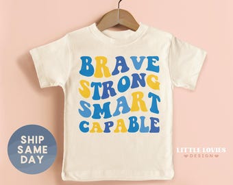 May include: Cream-coloured t-shirt with the words "BRAVE STRONG SMART CAPABLE" in blue and yellow bubble letters. The shirt is hanging on a wooden hanger against a pink background. A blue circle with the words "SHIP SAME DAY" is on the left.