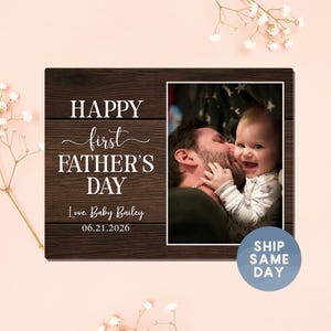 May include: A dark brown wooden picture frame with the text "HAPPY first FATHER'S DAY" in white. The frame includes a photo of a father kissing a baby. The text "Love, Baby Bailey 06.21.2026" is also visible.