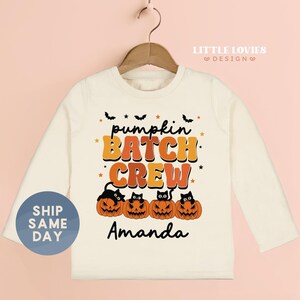 May include: Cream-colored long-sleeved shirt with the text "pumpkin BATCH CREW" in orange and brown. Below the text are four jack-o'-lanterns with black cat faces. The name "Amanda" is printed below the pumpkins. A blue circle with the text "SHIP SAME DAY" is on the left.