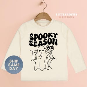 A white long-sleeved t-shirt with a graphic design featuring a ghost character and the text "SPOKEY SEASON BOO" printed on it.