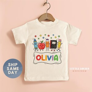 May include: A cream-colored short-sleeve t-shirt featuring a graphic of a blue crayon, a red apple, an orange glue stick, a black composition notebook, and a yellow pencil with faces, plus the name OLIVIA in colorful letters on a lined background.