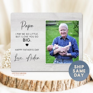 May include: A wooden photo frame with a picture of a man holding a baby. The frame has the words "Papa, I may be so little, but I love you so big. Happy Father's Day, Love, Aiden" written on it. The frame is on a wooden surface.