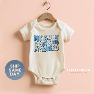 May include: A white baby onesie with the text "MY AUNT IS GETTING MARRIED" in blue bubble letters. The onesie is hanging on a wooden hanger against a light pink background. A blue circle with the text "SHIP SAME DAY" is on the left.