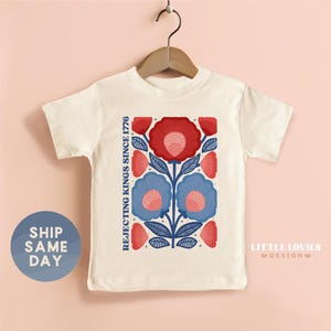 May include: Cream-colored t-shirt featuring a floral design in red and blue, with the text "Rejecting Kings Since 1776" on the left side. The shirt is hanging on a wooden hanger.