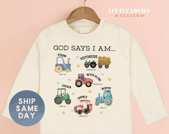 May include: Cream-colored long-sleeved shirt with a colorful tractor graphic and the text "GOD SAYS I AM...". The shirt features various tractor illustrations and inspirational words. A blue circle with the text "SHIP SAME DAY" is on the left.