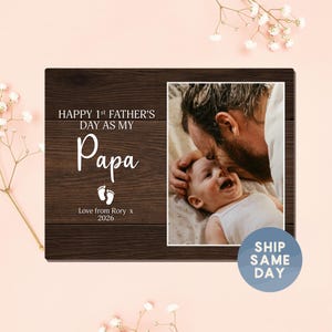 May include: A wooden photo frame with a picture of a father and baby. The left side of the frame has the text "HAPPY 1st FATHER'S DAY AS MY Papa" and "Love from Rory x 2026" in white. The frame is decorated with small white flowers.