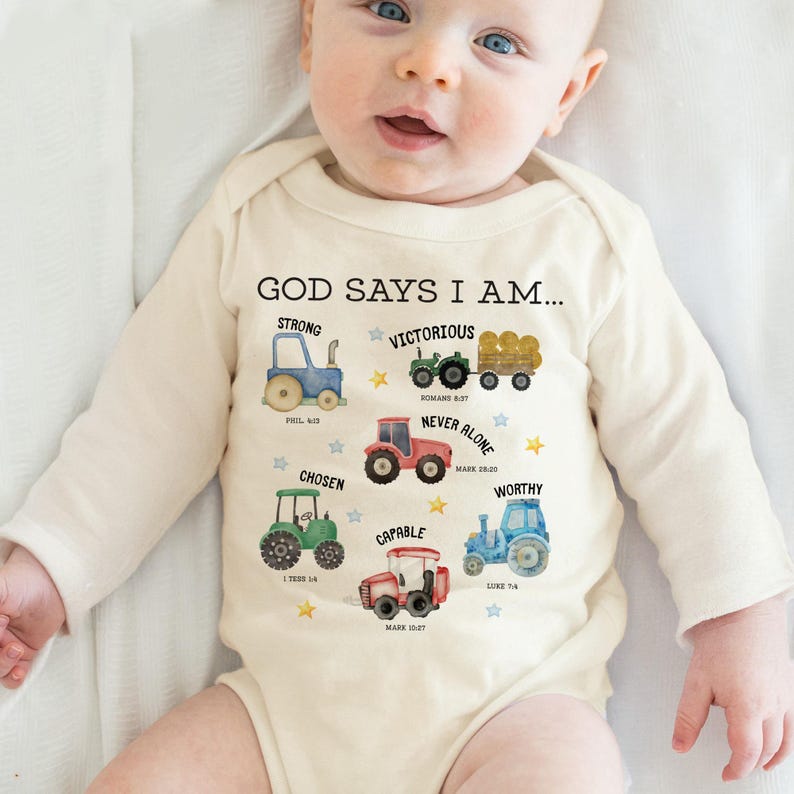 A baby wearing a white onesie with illustrations of various farm vehicles and the text "God says I am..."