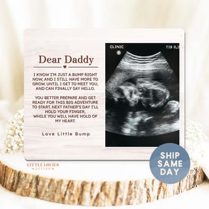 May include: A wooden photo frame with the text "Dear Daddy" and a sonogram image. The frame includes a message from the baby, expressing anticipation to meet their father. The frame is on a wooden surface.