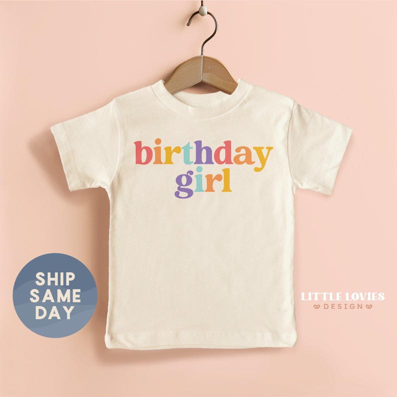 A white t-shirt with the text "BIRTHDAY GIRL" printed on it, hanging on a wooden hanger against a pink background.