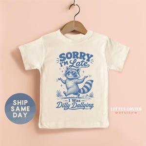 May include: A cream-colored short-sleeve t-shirt featuring a blue graphic of a raccoon wearing sunglasses. The text reads SORRY I'M LATE I WAS DILLY DALLYING. The shirt is displayed on a wooden hanger against a solid light pink background.