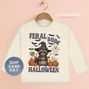 May include: Off-white long-sleeve shirt featuring a Halloween-themed graphic. The design includes a raccoon wearing a witch hat and cape, surrounded by pumpkins, bats, and the text "FERAL DUDE HALLOWEEN".