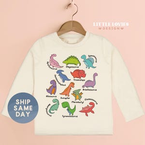 A white long-sleeved t-shirt with a colorful graphic design of various dinosaur illustrations, including a pink T-Rex, green Stegosaurus, purple Spinosaurus, and others.