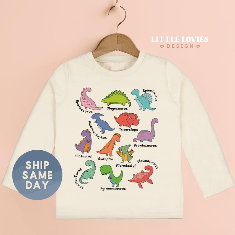 A white long-sleeved t-shirt with a colorful graphic design of various dinosaur illustrations, including a pink T-Rex, green Stegosaurus, purple Spinosaurus, and others.