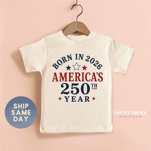 May include: A cream-colored short-sleeve t-shirt featuring the text BORN IN 2026 AMERICA'S 250TH YEAR in navy blue and red lettering. The design includes three small stars in navy, white, and red positioned above the red text.