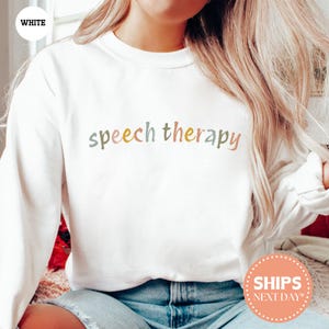 May include: White sweatshirt with the words "speech therapy" in pastel colors across the chest. The sweatshirt is a crew neck style and appears to be made of a soft material. The text is in a playful, handwritten font.