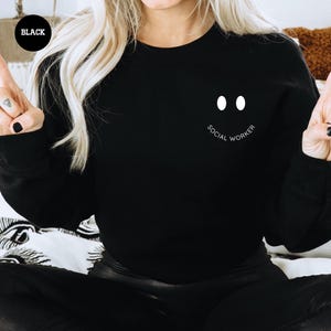 a woman sitting on a bed wearing a black sweatshirt with a smiley face drawn on