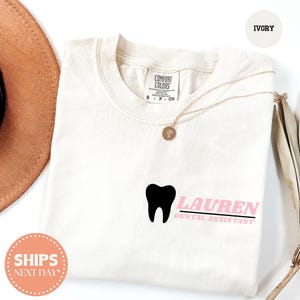 May include: An ivory short-sleeve t-shirt featuring a black tooth graphic and the text "LAUREN DENTAL ASSISTANT" in pink. The shirt has a crew neckline and a Comfort Colors brand label on the inside collar. A gold-toned necklace rests on the fabric.