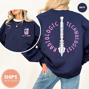 May include: A navy blue crewneck sweatshirt featuring a white graphic of a human spine on the back, surrounded by the pink text RADIOLOGIC TECHNOLOGIST. The front left chest displays a small pink graphic of a drink with the text AJ. R.T. (ARRT).
