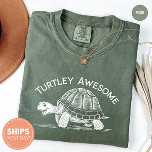 a green t-shirt with a turtle graphic and the text "TURTLEY AWESOME" printed on it. The t-shirt is laid out on a white surface, with a brown hat and some leaves visible in the background.