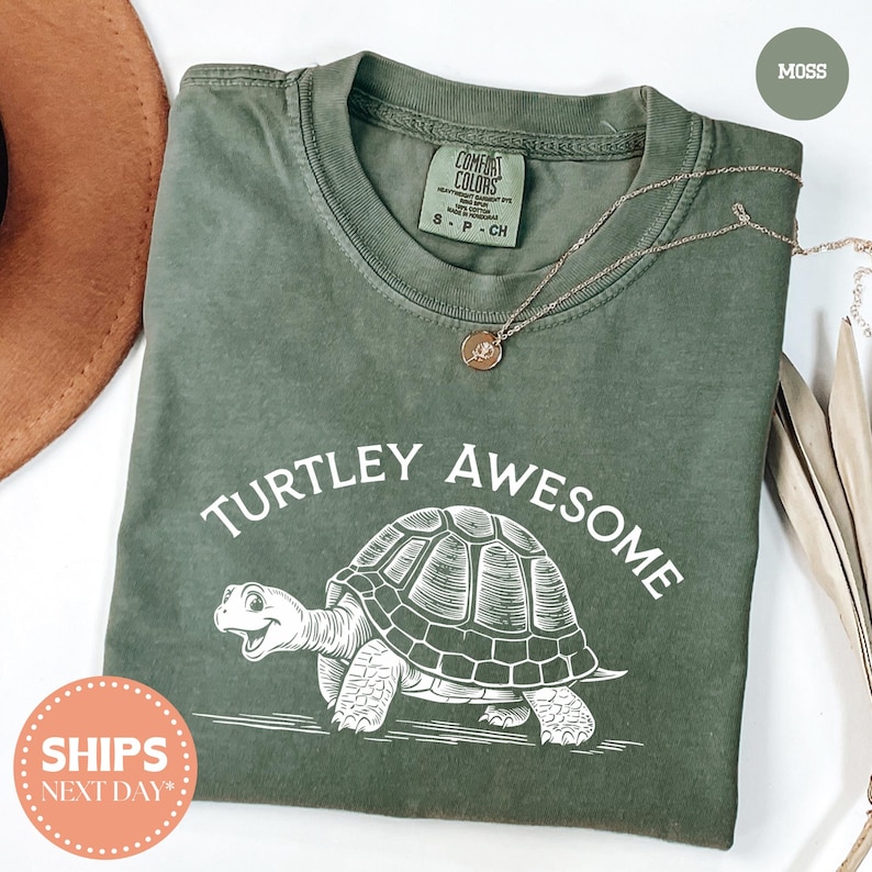 a green t-shirt with a turtle graphic and the text "TURTLEY AWESOME" printed on it. The t-shirt is laid out on a white surface, with a brown hat and some leaves visible in the background.