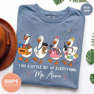 May include: A blue jean colored t-shirt featuring a graphic of four white geese dressed as teachers. The geese carry items like a snack box, coffee, school supplies, and a clipboard. The text reads I DO A LITTLE BIT OF EVERYTHING Ms. Anna.