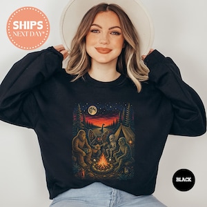 May include: A black long-sleeve sweatshirt featuring a graphic print of a campfire scene in a forest at night. The illustration depicts Bigfoot, a moth-like creature playing a guitar, an alien, and a swamp creature sitting around a fire.