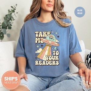 May include: A blue jean colored t-shirt featuring a graphic print of a flying saucer with an alien wearing a cowboy hat inside. The text reads TAKE ME TO YOUR READERS in a retro font, surrounded by stars, clouds, and lightning bolt accents.