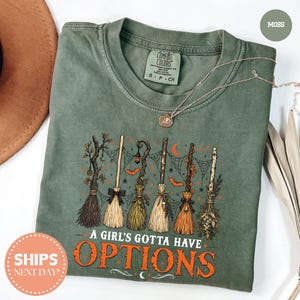 May include: A moss green crew neck t-shirt featuring a graphic of six different witch brooms, spiderwebs, bats, and crescent moons. The text "A GIRL'S GOTTA HAVE OPTIONS" is printed in orange below the brooms. A thin gold chain necklace rests on the shirt.