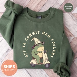 May include: Military green sweatshirt with the phrase "OFF TO COMMIT MOM FOOLERY" in a circular design. The graphic features a frog wearing a wizard hat and riding a cream-colored scooter. The sweatshirt has long sleeves.