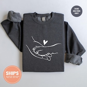 May include: Dark heather grey sweatshirt with a white graphic of two hands holding each other, with a heart above. The sweatshirt has a crew neck and long sleeves with ribbed cuffs. The text "DARK HEATHER GREY" is in a circle.