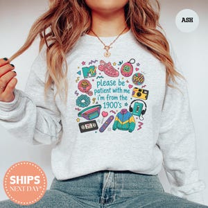 May include: An ash gray sweatshirt featuring a colorful graphic print of 1990s nostalgia items including an MTV logo, a jelly shoe, a digital pet, a fanny pack, a cassette tape, and a windbreaker, with the text please be patient with me i'm from the 1900's.