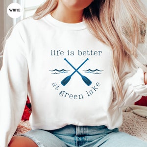 a woman sitting on a bed wearing a white sweatshirt that says life is better at