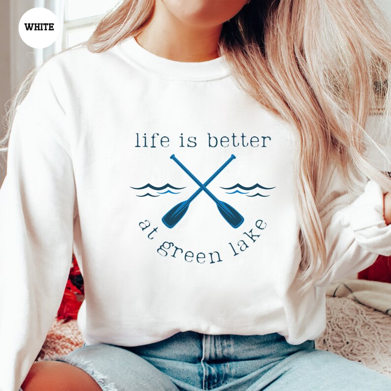 a woman sitting on a bed wearing a white sweatshirt that says life is better at