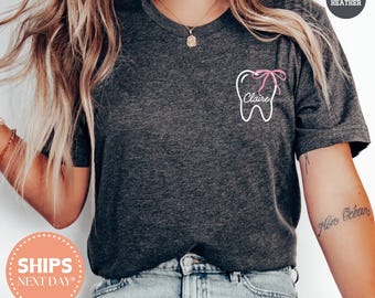 May include: Dark grey t-shirt with a white tooth outline and a pink bow, the name "Claire" is written in cursive. The shirt has a round neck and short sleeves. The text "DARK HEATHER" is in a circle in the upper right corner.