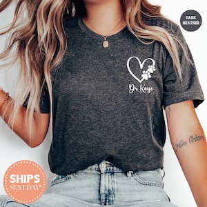 May include: Dark heather gray t-shirt with a white heart and paw print design. The text "Dr. Kaye" is written below the heart. The shirt has a crew neck and short sleeves.