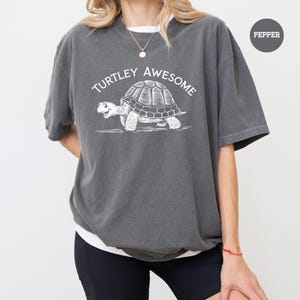 A person wearing a gray t-shirt with a graphic of a turtle and the text "TURTLEY AWESOME" printed on it.