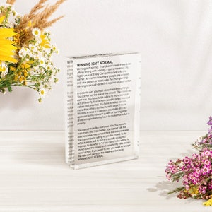Könnte beinhalten: Ein weißer Acrylblock mit dem Text "WINNING ISN'T NORMAL" in Schwarz gedruckt. Der Text ist ein Zitat über die Natur des Gewinnnens und wie es ungewöhnliche Aktionen und die Bereitschaft erfordert, aus der Masse herauszustechen.